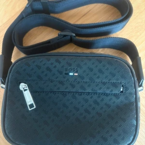 Svart axelväska från BOSS med mönster - Snygg svart axelväska från BOSS med geometriskt mönster och silverfärgad dragkedja. Väskan har justerbar rem och ett rymligt fack med innerficka. Materialet är skinn och polyester, perfekt för dig som gillar stilrena accessoarer med coola detaljer.