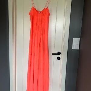Långklänning med smala axelband - Säljer en aprikos långklänning med smala axelband. Klänningen har volangdetaljer vid axlarna och knytband i midjan för en snygg siluett. Dragkedja i sidan. 