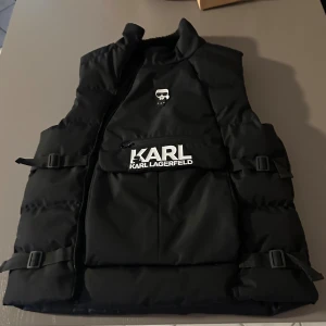 Svart dunväst från Karl Lagerfeld - Svart dunväst från Karl Lagerfeld med coolt tryck på bröstet och stor logotyp på fickan. Västen har justerbara spännen på sidorna, hög krage och quiltad design. Materialet är polyester och insidan är mjukt fodrad. Perfekt för lager-på-lager och streetstyle.