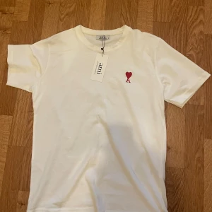 Vit t-shirt från Ami med rött hjärta - Vit t-shirt från Ami Alexandre Mattiussi i mjuk bomull. Klassisk passform med korta ärmar och rund hals. På bröstet finns ett broderat rött hjärta med bokstaven A. Perfekt basic med en snygg detalj framtill.