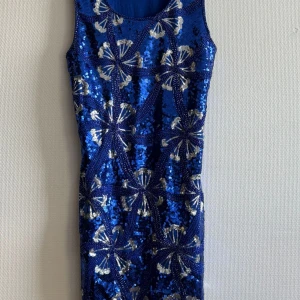 Blå paljettklänning med mönster - Snygg blå ärmlös kortklänning täckt av glittriga paljetter och silverfärgat mönster framtill. Klänningen har en figurnära passform och är perfekt för dig som vill sticka ut. Baksidan är enfärgad blå i mjukt tyg.