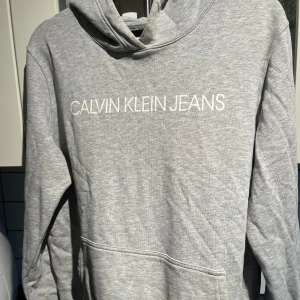Grå hoodie från Calvin Klein Jeans - Säljer en stilren grå hoodie från Calvin Klein Jeans med vit logga framtill. Tröjan har huva, magficka och långa ärmar. Perfekt för en chill och avslappnad look.