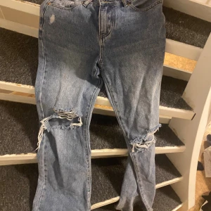 Blå slitna jeans med hål på knäna - Säljer ett par blå jeans med coola slitningar och stora hål på båda knäna. De har en klassisk femficksdesign och bootcut.