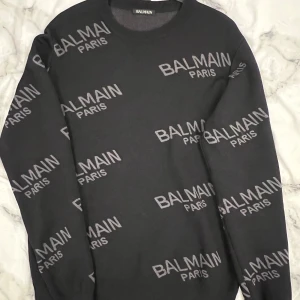 Svart stickad tröja Balmain Paris - Snygg svart stickad tröja från Balmain Paris med grå logotyp tryckt över hela tröjan. Rund halsringning och långa ärmar ger en clean och modern look. Perfekt för dig som gillar exklusiva streetwear-vibbar och vill sticka ut.