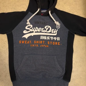 Blå och svart Superdry hoodie M - Snygg hoodie från Superdry i blått och svart med vit och orange text framtill. Tröjan har huva med vita snören och känguruficka. Perfekt för dig som gillar streetwear och vill ha en bekväm och stilren look.