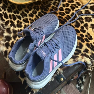 Adidas sneakers lila/blå med rosa ränder - Blå/lila sneakers från Adidas med klassiska rosa ränder på sidorna. Ovandelen är i mesh för bra andningsförmåga och skorna har snörning samt vit sula. Perfekta för dig som gillar sportig stil eller vill ha bekväma skor till vardagen.