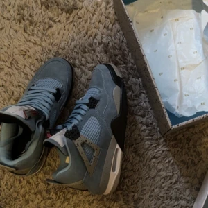 Nike Air Jordan 4 Retro University Blue - Snygga Nike Air Jordan 4 Retro i University Blue med grå och svarta detaljer. Skorna har en klassisk siluett, meshpaneler och snörning. Ovandelen är i mocka och sulan är vit med svarta inslag. Perfekta för dig som vill sticka ut med stilrena sneakers.