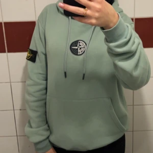 Grön hoodie från Stone Island - Snygg ljusgrön hoodie från Stone Island med klassisk patch på ärmen och logga på bröstet. Tröjan har huva med snörning och en stor magficka. Perfekt för dig som gillar streetwear och vill ha en chill look.