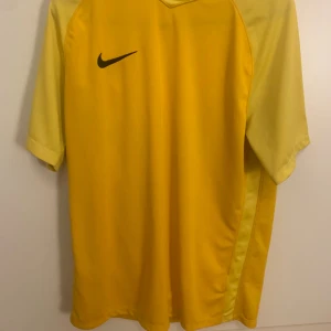 Gul Nike Dri-Fit fotbollströja L - Gul fotbollströja från Nike i Dri-Fit-material som andas. Tröjan har korta ärmar och en klassisk passform med svart Nike-logga på bröstet. Perfekt för träning eller match, tillverkad i Thailand.