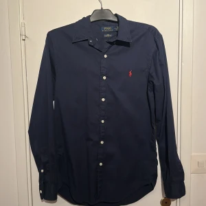 Mörkblå skjorta Polo Ralph Lauren - Snygg mörkblå skjorta från Polo Ralph Lauren i slim fit med klassisk krage och vita knappar. Materialet är lätt bomullstwill och har den ikoniska röda Polo-loggan broderad på bröstet. Skjortan är använd fåtal gånger är som ny. Nypris 1600 mitt pris 600