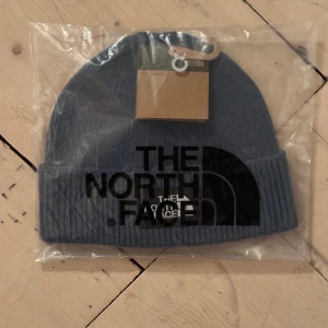 Blågrön mössa från The North Face - Snygg blå ribbstickad mössa från The North Face med stor svart logga framtill. Klassisk uppvikt kant och mjukt material som håller dig varm. Perfekt för dig som vill ha en clean och stilren look under kalla dagar.