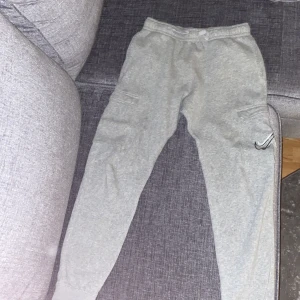 Grå cargobyxor från Nike, storlek L - Säljer ett par grå cargobyxor från Nike i storlek L. Byxorna har elastisk midja, cargofickor på sidorna och flera snygga Nike-loggor tryckta på ena benet. Tillverkade i mjukt bomullsmaterial för en skön och avslappnad look.