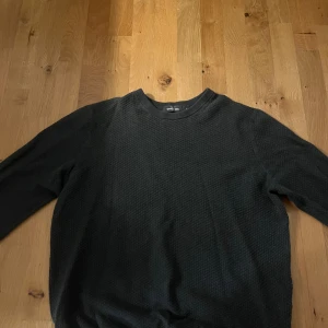 Svart stickad tröja från Dressmann - Svart stickad tröja från Dressmann i storlek M. Tröjan har en diskret struktur i materialet, rund halsringning och långa ärmar. Perfekt för dig som gillar en enkel och stilren look.