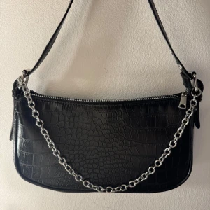 Svart axelväska med kedja - Snygg svart axelväska med krokodilpräglat mönster och silverfärgad kedjedetalj framtill. Väskan har dragkedja upptill och en stilren, rektangulär form. Perfekt för att ge din outfit en cool vibe.