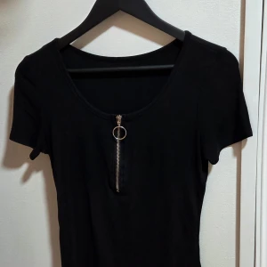 Svart ribbad topp med dragkedja - Svart ribbad topp med rund halsringning och korta ärmar. Framtill finns en silverfärgad dragkedja med ring som detalj, vilket ger en cool vibe. Materialet är stretchigt och sitter tight mot kroppen. Perfekt för dig som gillar minimalistisk stil med en twist.