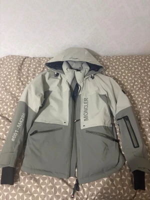 Moncler grenoble - Säljer en grå och ljusgrå vindjacka från Moncler med huva och stora tryck på både fram- och baksida. Jackan har dragkedjor på fickorna, muddar vid ärmslut och coola detaljer som #001-M4286 på ärmen och 'HIGH PERFORMANCE' på ryggen. STORLEK S/M