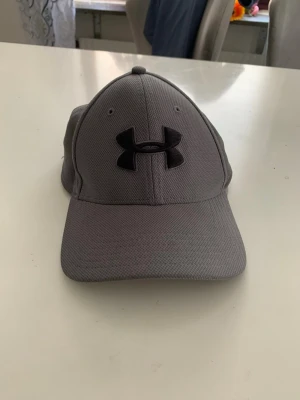 Grå keps från Under Armour - Snygg grå keps från Under Armour med svart logga framtill. Perfekt för sport och vardag. Kepsen har en klassisk design med böjd skärm och ventilationshål för extra komfort.