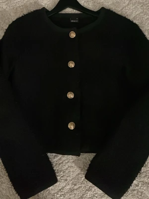 Svart kofta med guldknappar Gina Tricot - Säljer en svart kofta från Gina Tricot med fem dekorativa guldknappar framtill. Koftan har en mjuk, lite lurvig yta och rund halsringning. Perfekt till lager på lager och ger en chic touch till din outfit.