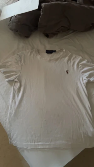 Vit t-shirt från Polo Ralph Lauren - Vit t-shirt från Polo Ralph Lauren med klassisk rund hals och korta ärmar. Diskret broderad logga på bröstet. Mjuk och skön bomull som passar perfekt till jeans eller shorts. Enkel och stilren design för en clean look.