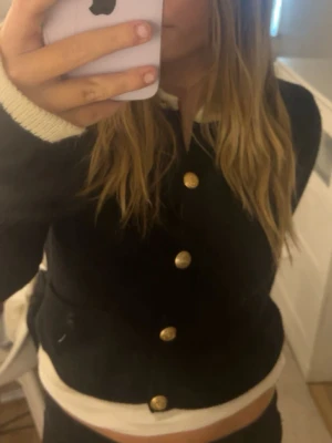 Svart kofta med guldknappar - Säljer en svart kofta med snygga guldknappar framtill. Den är lite tjockare, nästan som en cardigan.  Koftan har lång ärm och vita detaljer vid ärmslut och krage. Perfekt för dig som vill ha en klassisk men ändå trendig look.