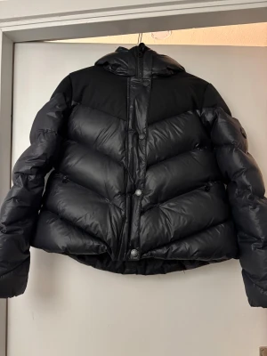 Ella&il Jennie down jacket  - Säljer en svart pufferjacka med huva och snygga sneda sömmar. Jackan har både dragkedja och knappar framtill, samt en lite croppad passform. Perfekt för dig som vill ha en varm och stilren jacka med modern look. Som ny skick förutom ett litet hål vid dragkedjan. ”Bältet” till den saknas. Nypris 5599kr. Skriv vid funderingar💞