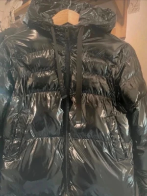 Svart pufferjacka från Guess - Säljer en glansig svart pufferjacka från Guess med huva och breda snören. Jackan har dragkedja framtill och är tillverkad i återvunnen polyester. Perfekt för kalla dagar och riktigt snygg streetstil.