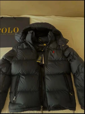 Svart pufferjacka från Polo Ralph Lauren - Säljer en svart pufferjacka från Polo Ralph Lauren med huva och dragkedjor. Jackan har det klassiska röda Polo-logot broderat på bröstet och två sidofickor med dragkedja. Perfekt för kalla dagar och riktigt snygg till streetwear-stilen.