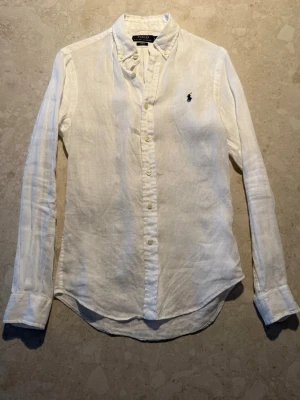 Vit Polo ralph lauren linneskjorta - Snygg vit skjorta i linne från Polo Ralph Lauren med klassisk button-down krage och broderad marinblå logga på bröstet. Slim fit-modell med långa ärmar och knappar framtill. Perfekt för dig som gillar stilrena och fräscha plagg. Pris 579kr! Storlek xs slimfit!