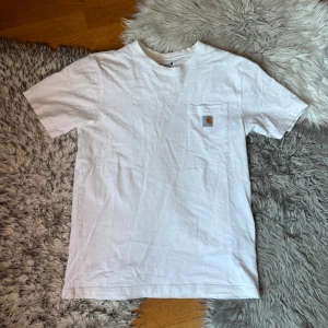 Vit Carhartt t-shirt med bröstficka - Vit t-shirt från Carhartt i relaxed fit med klassisk bröstficka och Carhartt-logga på fickan. Tillverkad i mjuk bomull och har en enkel, clean look med rund hals och korta ärmar. Perfekt till jeans eller shorts.