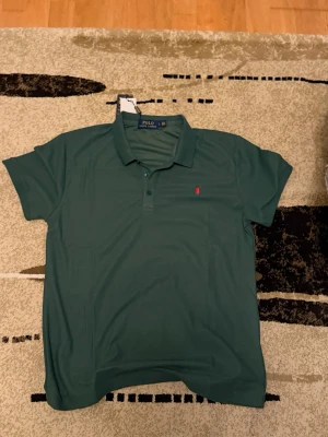 Grön pikétröja från Polo Ralph Lauren - Snygg mörkgrön pikétröja från Polo Ralph Lauren med klassisk krage och knappar framtill. Tröjan har korta ärmar och den ikoniska röda logotypen broderad på bröstet. Tillverkad i mjuk bomull som känns skön mot huden.