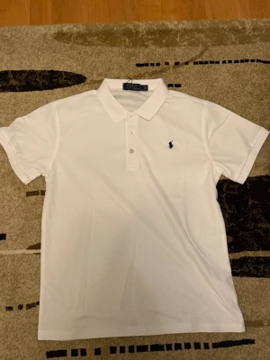 Vit pikétröja från Polo Ralph Lauren - Klassisk vit pikétröja från Polo Ralph Lauren med korta ärmar och krage. Tröjan har två knappar framtill och den ikoniska broderade loggan i svart på bröstet. Tillverkad i mjuk bomull som känns skön mot huden. Perfekt till jeans eller shorts.