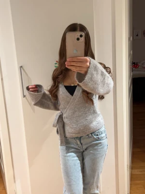 Grå omlottkofta från Gina  - Mysig grå omlottkofta från gina med knyt i midjan och v-ringad hals. Koftan är stickad och har långa, lite vidare ärmar. 