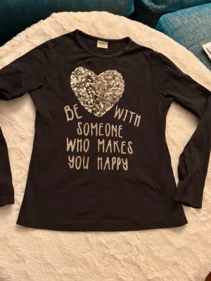 Svart långärmad topp Zara Girls - Svart långärmad topp från Zara Girls med ett stort hjärta i glittriga paljetter på bröstet och texten 'Be with someone who makes you happy' framtill. Mjuk och skön bomull, perfekt till vardags för dig som gillar lite extra bling.