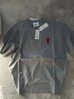 Grå t-shirt från Ami Paris med rött hjärta - Snygg grå t-shirt från Ami Paris med klassisk rund hals och ett broderat rött hjärta med bokstaven A på bröstet. Tillverkad i mjuk bomull för skön känsla och enkel stil. Perfekt till jeans eller shorts för en clean look.
