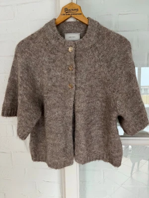 Beige stickad kofta från Neo Noir - Mysig beige stickad kofta från Neo Noir med kort ärm och snygga guldknappar framtill. Koftan har en fluffig känsla och rund halsringning, perfekt för lager-på-lager under hösten eller vintern.