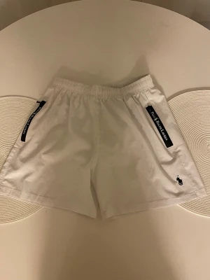 Vita badshorts från Polo Ralph Lauren - Säljer ett par fräscha vita shorts från Polo Ralph Lauren med elastisk midja och svarta detaljer vid fickorna där märkesnamnet står. Klassisk logga broderad på benet. Perfekta för sommaren och riktigt clean look.
