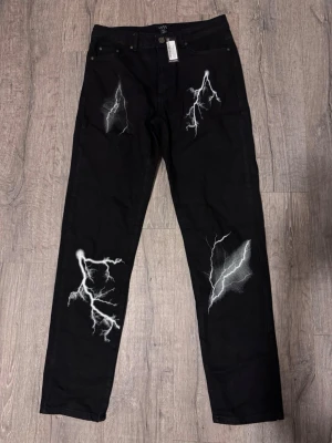 ⚡️Black Lightning Print Jeans / Trousers ⚡ -  Stand out with these black lightning bolt print trousers – bold, edgy and perfect for streetwear or alternative fashion lovers. Features an all-over white lightning design on a black base for a striking look.  📏 Length: approx. 117 cm 🎨 Colour: Black & White 👖 Style: Straight leg / casual fit ✨ Condition: Very good 🔥 Vibe: Y2K, grunge, streetwear, alt  Perfect for pairing with hoodies, crop tops, boots or sneakers for a statement outfit.