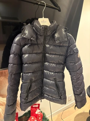 Moncler Bady dunjacka mörkblå - Intressekoll på min snygg Moncler Bady dunjacka i mörkblå färg med glansig finish. Jackan har huva, två framfickor med dragkedja och Moncler-logga på ärmen. Puffad design och hel dragkedja framtill. Finns tecken på användning på insidan när man kollar på den stora ettiken på sidan men inget man tänker på och kan kanske lagas. Kvittot och prislappen finns kvar.