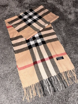 Rutig cashmerehalsduk från Burberry - Klassisk beige halsduk från Burberry i 100% kashmir. Den har det ikoniska rutiga mönstret i svart, vitt och rött samt fransar i ändarna. Perfekt accessoar för att lyfta din outfit med en tidlös brittisk touch.