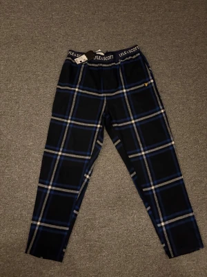 Rutig pyjamas byxa från Lyle & Scott - Säljer ett par svarta och blåa pyjamas byxor från Lyle & Scott med stort rutmönster och resårmidja med logga. Byxorna har en gul broderad logga på benet och är gjorda i mjukt bomullstyg. Perfekta för chill dagar hemma.