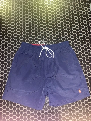 Marinblå badshorts från Polo Ralph Lauren - Säljer ett par marinblå badshorts från Polo Ralph Lauren med vit snörning i midjan och klassisk röd logga broderad på benet. Dem passar mer som S/M kolla måtten på bilden Shortsen har elastisk midja, sidofickor och en bakficka. Perfekta för stranden eller poolen.
