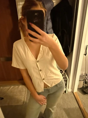 Kortärmad vit topp med knappar - Säljer en stilren vit topp med v-ringning och korta ärmar. Toppen har en avslappnad passform och knäpps med tre synliga knappar framtill. Perfekt till jeans eller kjol för en enkel och fräsch look.