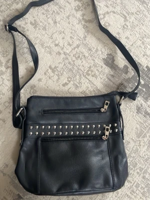 Svart axelväska med nitar - Säljer en svart axelväska i skinnimitation med justerbart axelband. Väskan har flera dragkedjefack och en rad med silvriga nitar framtill som ger en cool vibe. Perfekt storlek för att få plats med allt du behöver och enkel att matcha till olika outfits.