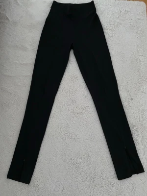 Svarta leggings med slits från Gina Tricot - Säljer ett par svarta leggings från Gina Tricot i storlek S. De har hög midja, tight passform och slits med dragkedja längst ner på benen. Materialet är stretchigt och bekvämt, perfekt till en snygg och clean look. Byxorna är mycket väl skötta som om de vore i ny skick.