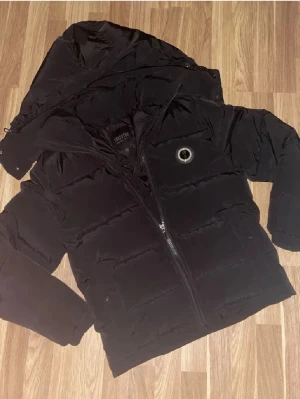 Svart pufferjacka Trapstar XS - Svart pufferjacka från Trapstar i storlek XS. Jackan har en stor huva, broderad Trapstar-logga på ryggen och dragkedja framtill. Tillverkad i 100% polyester med ett clean och stilrent utseende, perfekt för kalla dagar.