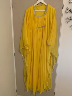 Gul långklänning med strassdetaljer - Flödande gul långklänning med glittrande strass runt fyrkantig ringning och under bysten. Vida ärmar och silverkantade avslut ger en extra lyxig känsla. Perfekt för dig som vill sticka ut med färg och elegans. Köpt i Dubai. Aldrig använd. Stl S, men passar även M. Arabisk design klänning. 