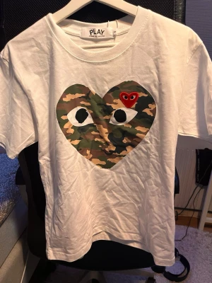 Vit Comme des Garçons Play t-shirt - Säljer en vit t-shirt från Comme des Garçons Play med ikoniskt hjärta i camouflage och ett litet rött hjärta med ögon på bröstet. Klassisk passform och rund halsringning. Perfekt statement-plagg för dig som gillar streetwear.
