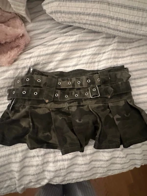 Camo minikjol med bälte - Säljer en kort camouflagekjol med bredt bälte och silverfärgade nitar. Kjolen har plisserad nederdel och dragkedja i sidan. Aldrig använd. Skulle nästan säga xs men storleken är s