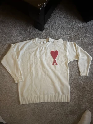 Vit stickad tröja från Ami - Säljer en vit stickad tröja från Ami Alexandre Mattiussi med röd hjärtdetalj och bokstaven A på bröstet. Tröjan har rund halsringning, ribbade muddar och är långärmad. Perfekt för dig som vill ha en clean men ändå statement look.