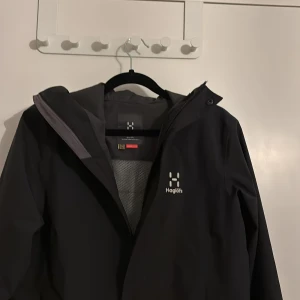 Svart regnjacka från Haglöfs med huva - Svart regnjacka från Haglöfs med minimalistisk design och justerbar huva. Jackan har dragkedja framtill, diskret logga på bröstet och dragsko nertill. Perfekt för blöta dagar och enkel att matcha med allt. Snygg och funktionell för dig som gillar outdoor-vibe.